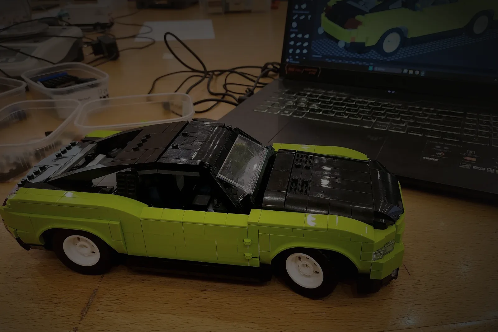 A lime green and black car model on the studio bench, with the same car open in the design software on the laptop directly behind it, its parts list down the side of the screen.