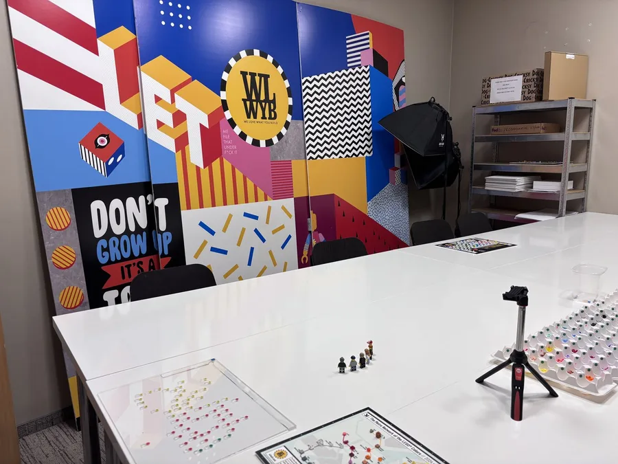 A studio table set up for photography, with a line of minifigures and colour-sorted trays, in front of a graphic wall reading LET, the WLWYB mark, and DON'T GROW UP, IT'S A TOY.