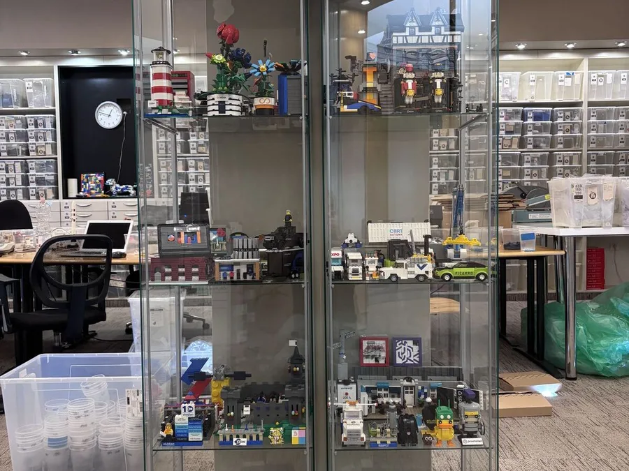 A tall glass display cabinet whose shelves hold rows of finished custom models built from LEGO® bricks, standing in front of the parts wall.