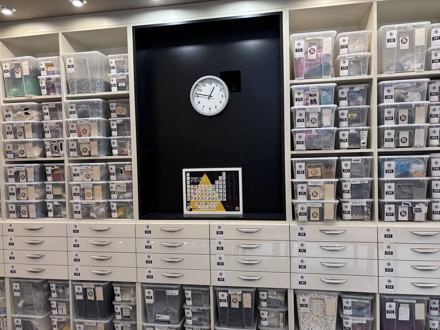 A wall of labelled storage bins and drawers holding sorted LEGO® parts by numbered ID, with a wall clock and a framed periodic table of LEGO® colours on the back panel.
