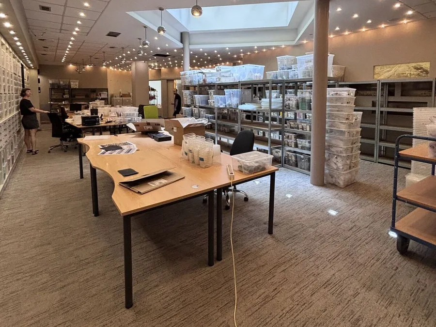A wide view of the studio floor: desks, a person at the parts wall, shelving stacked with storage bins, and a skylight overhead.