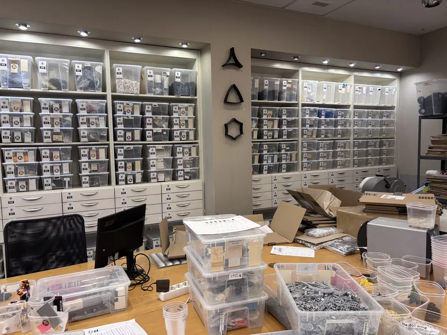 A work desk with a monitor and open parts trays, backed by floor-to-ceiling shelving of labelled storage bins.